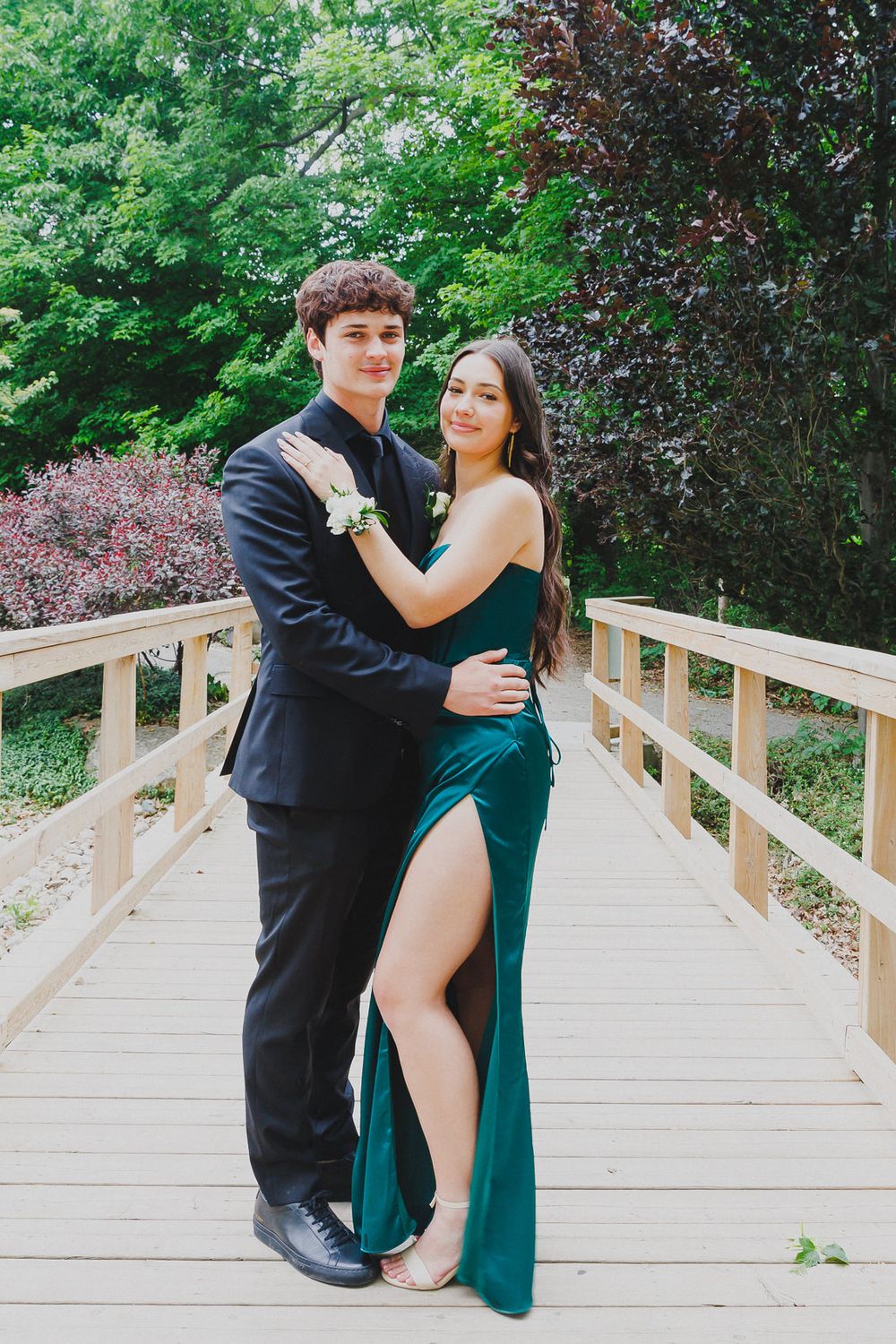 Formal couple posing on wooden bridge surrounded by lush spring foliage.
