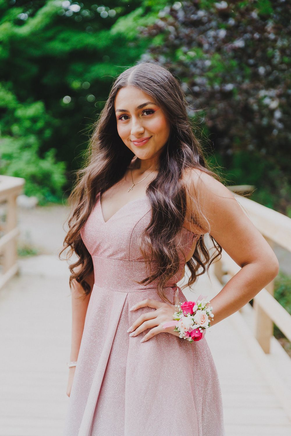 Elegant pink formal dress with floral wrist corsage photographed on outdoor wooden bridge.