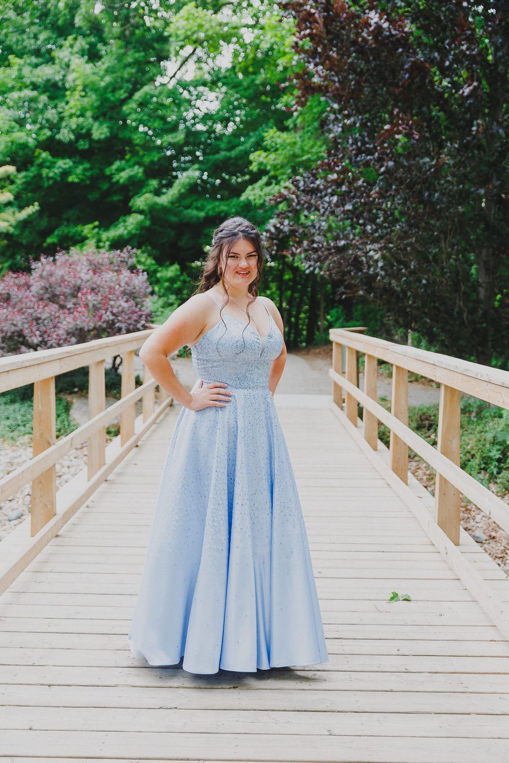 Person in light blue formal gown poses on wooden bridge with spring foliage backdrop.