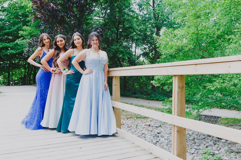 Four people in elegant prom dresses pose on wooden bridge with green foliage background.