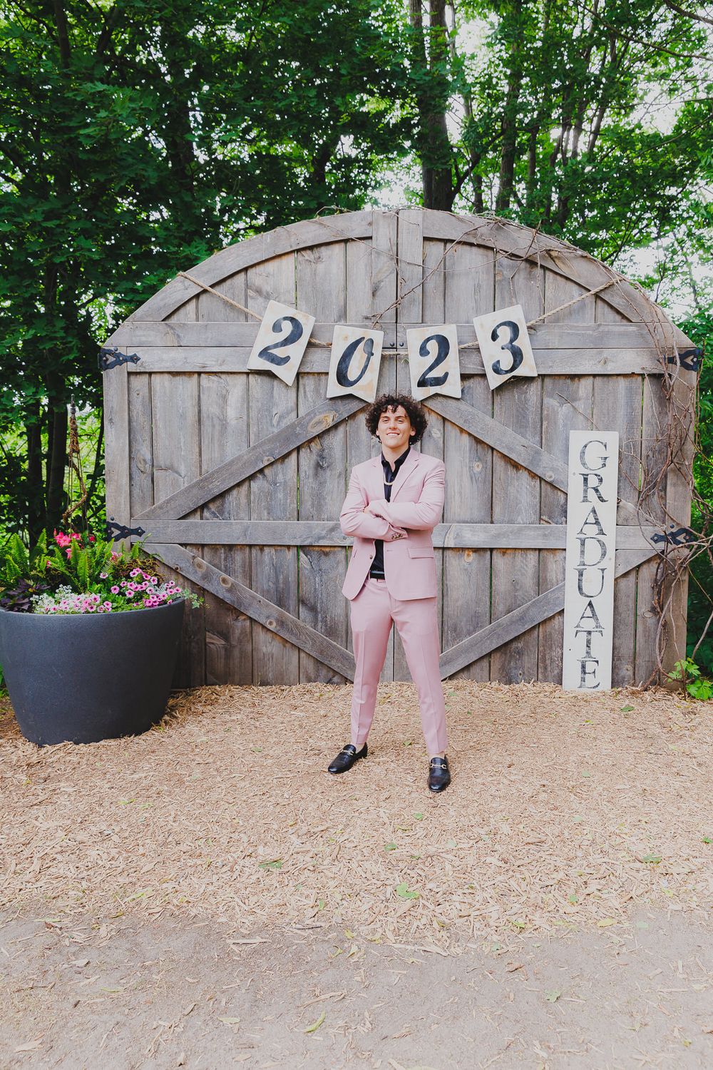 A graduate in a pink suit poses in front of a rustic wooden barn door with '2023' numbers displayed.