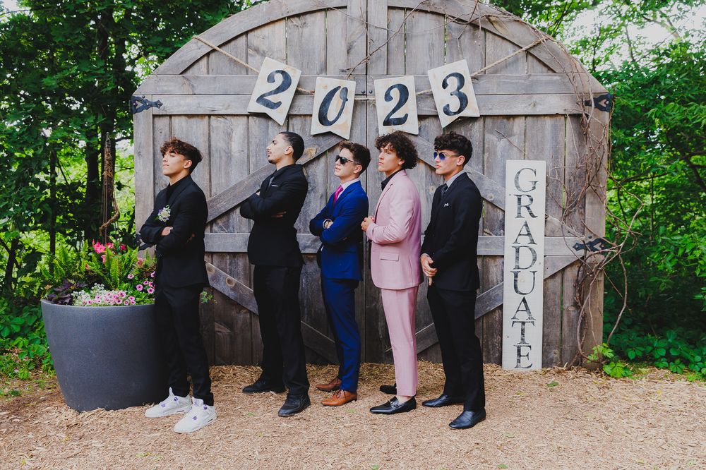 Five graduates pose together in formal attire against a rustic barn door decorated with '2023'.