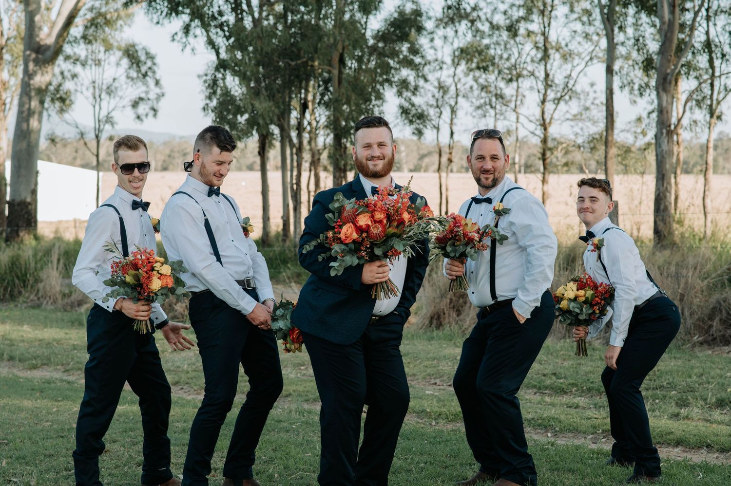 Groomsmen in suspenders and bow ties pose with orange flower bouquets against eucalyptus trees.