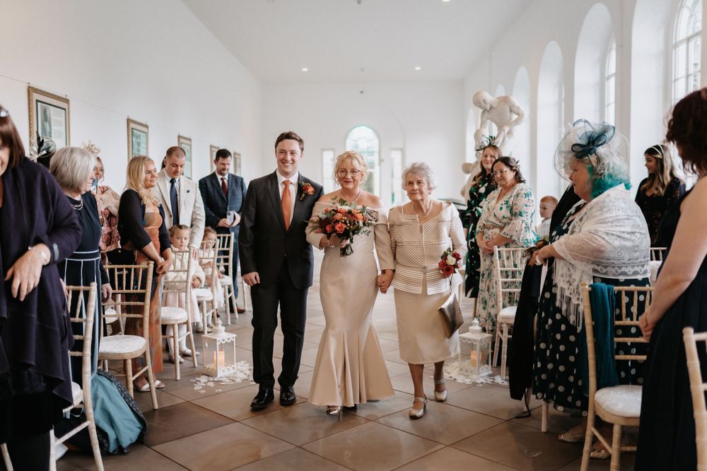 Wedding guests line aisle in bright indoor venue with arched windows at The Orangery Margam Park