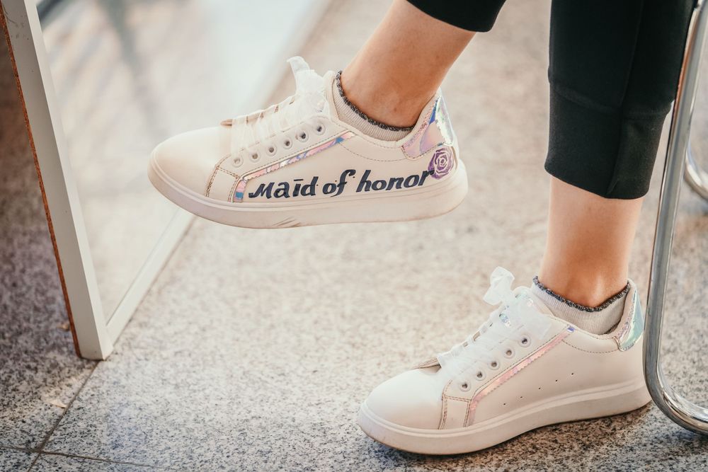 White sneakers with 'maid of honor' text written on the side against a light floor.