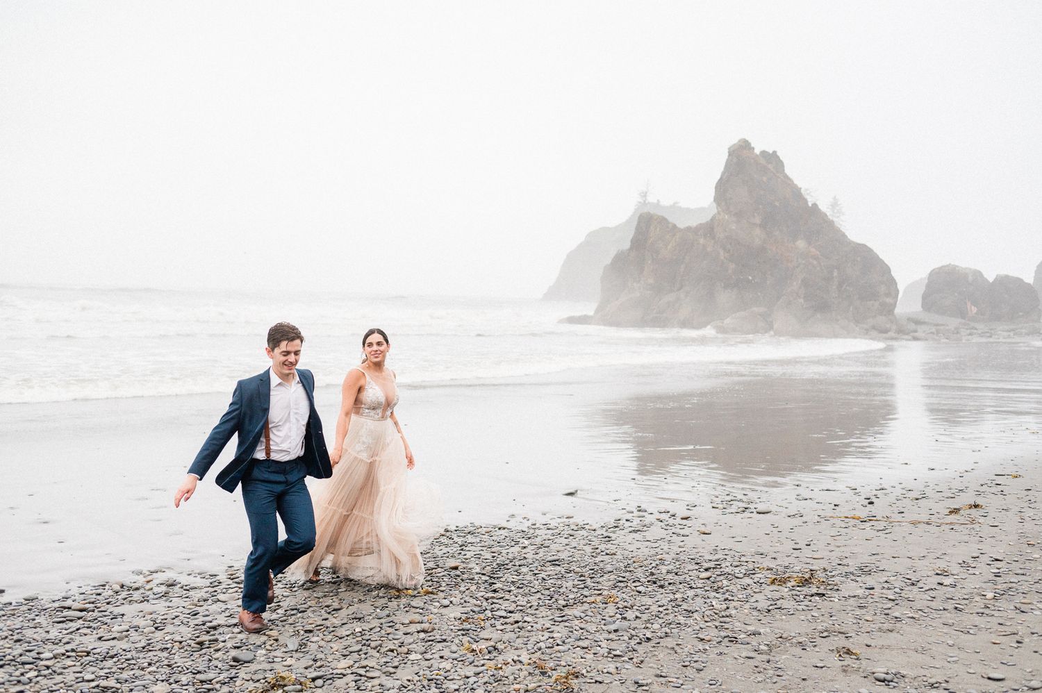 Bride and groom running along a foggy beach.