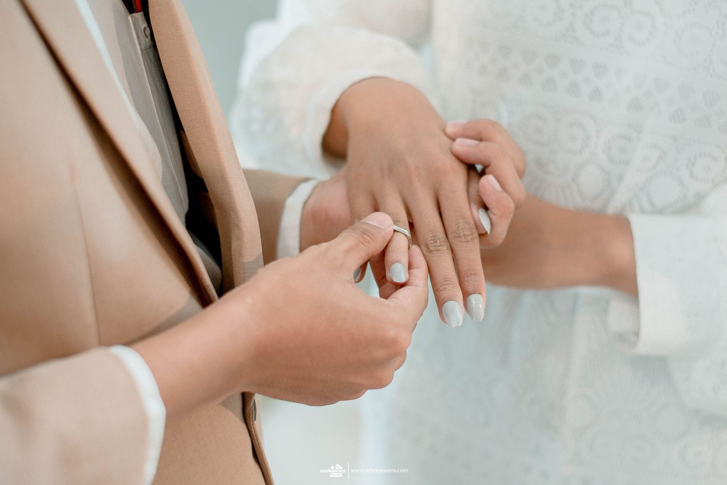Close-up of hands exchanging wedding rings against neutral-toned formal attire.