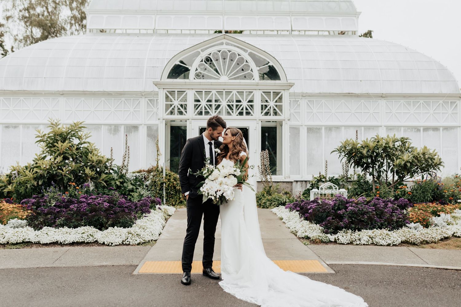 Elegant wedding photo in front of a white conservatory with beautiful flower gardens.