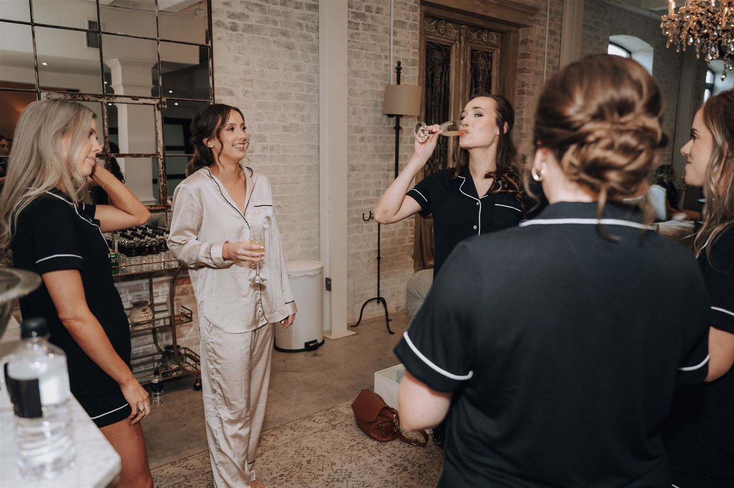 Group of people getting ready in a bridal suite with exposed brick walls and warm lighting.