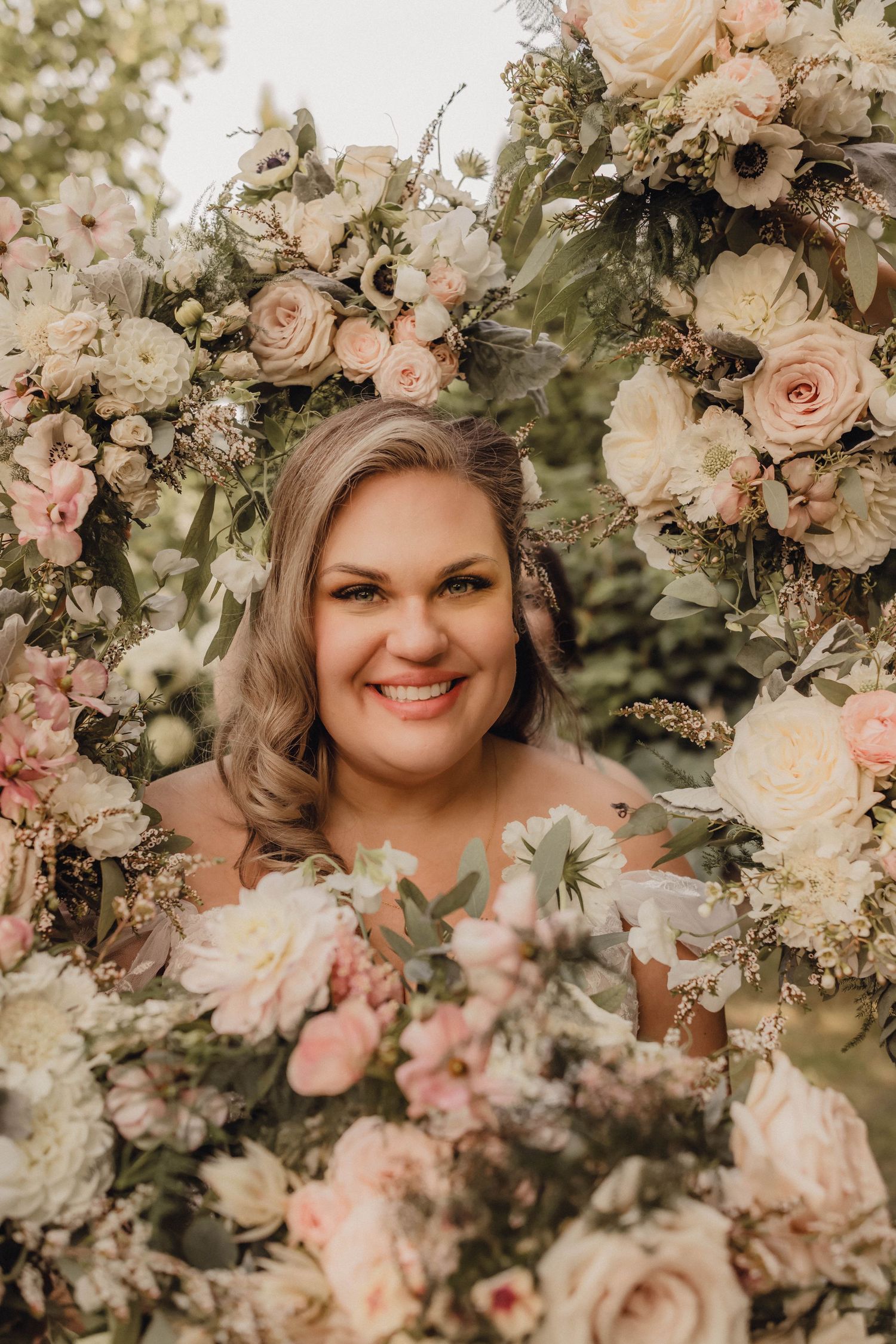Portrait of a smiling bride surrounded by soft floral arrangements, capturing the joy of the wedding day.