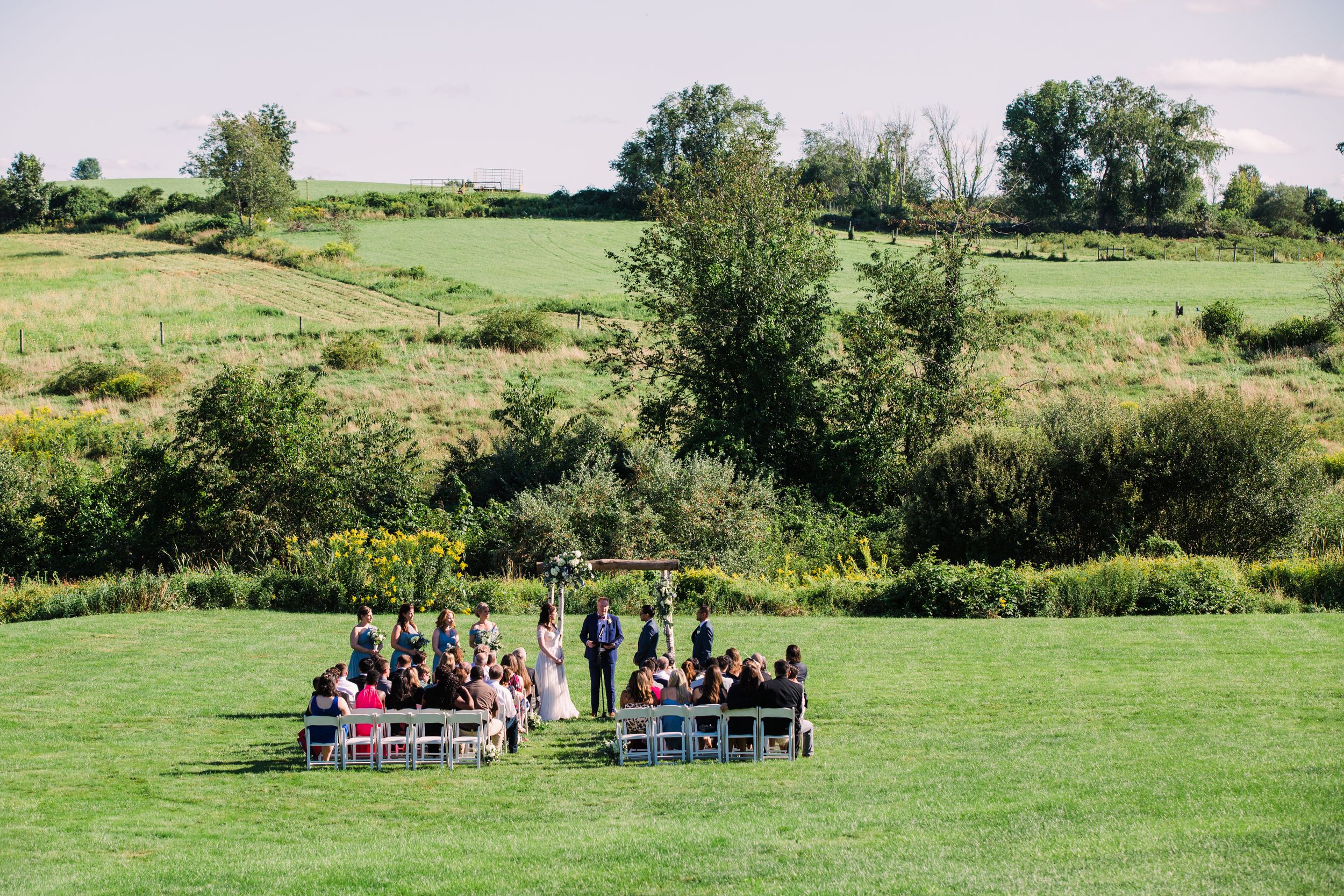 Intimate outdoor wedding ceremony setup in a lush green meadow with rolling hills in the background.