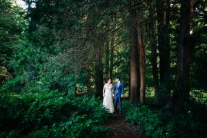 A couple in wedding attire walks through a lush forest path surrounded by green foliage.