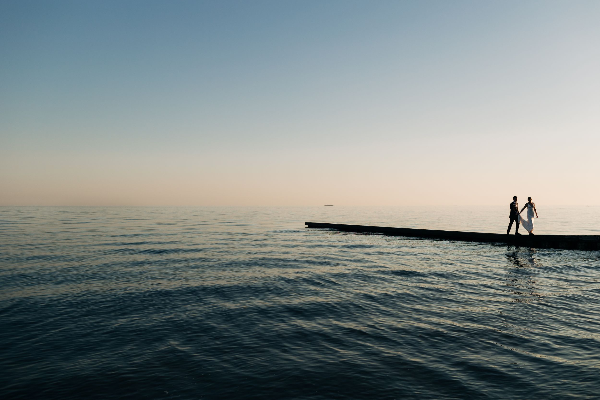 Two silhouettes stand on a pier at sunset overlooking calm ocean waters.