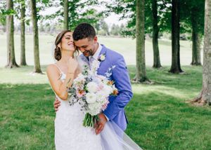 Wedding couple sharing intimate moment with white floral bouquet against tree-lined backdrop.