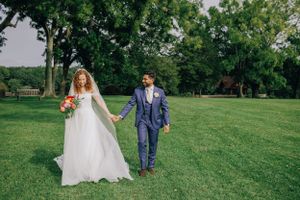 Couple in wedding attire walking hand in hand across a sunlit lawn with trees in background.