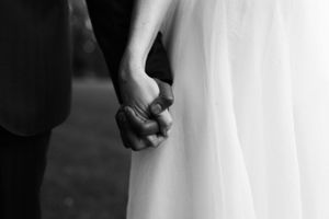 Black and white close-up of newlyweds holding hands, showing wedding dress detail.