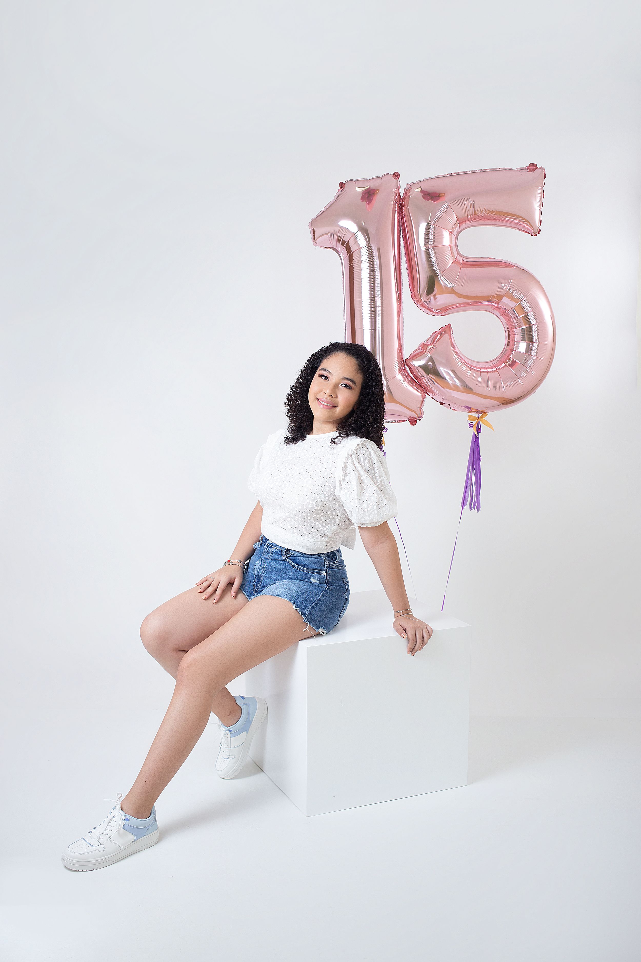 Festive quinceañera photoshoot with pink number balloons and white podium against white backdrop.