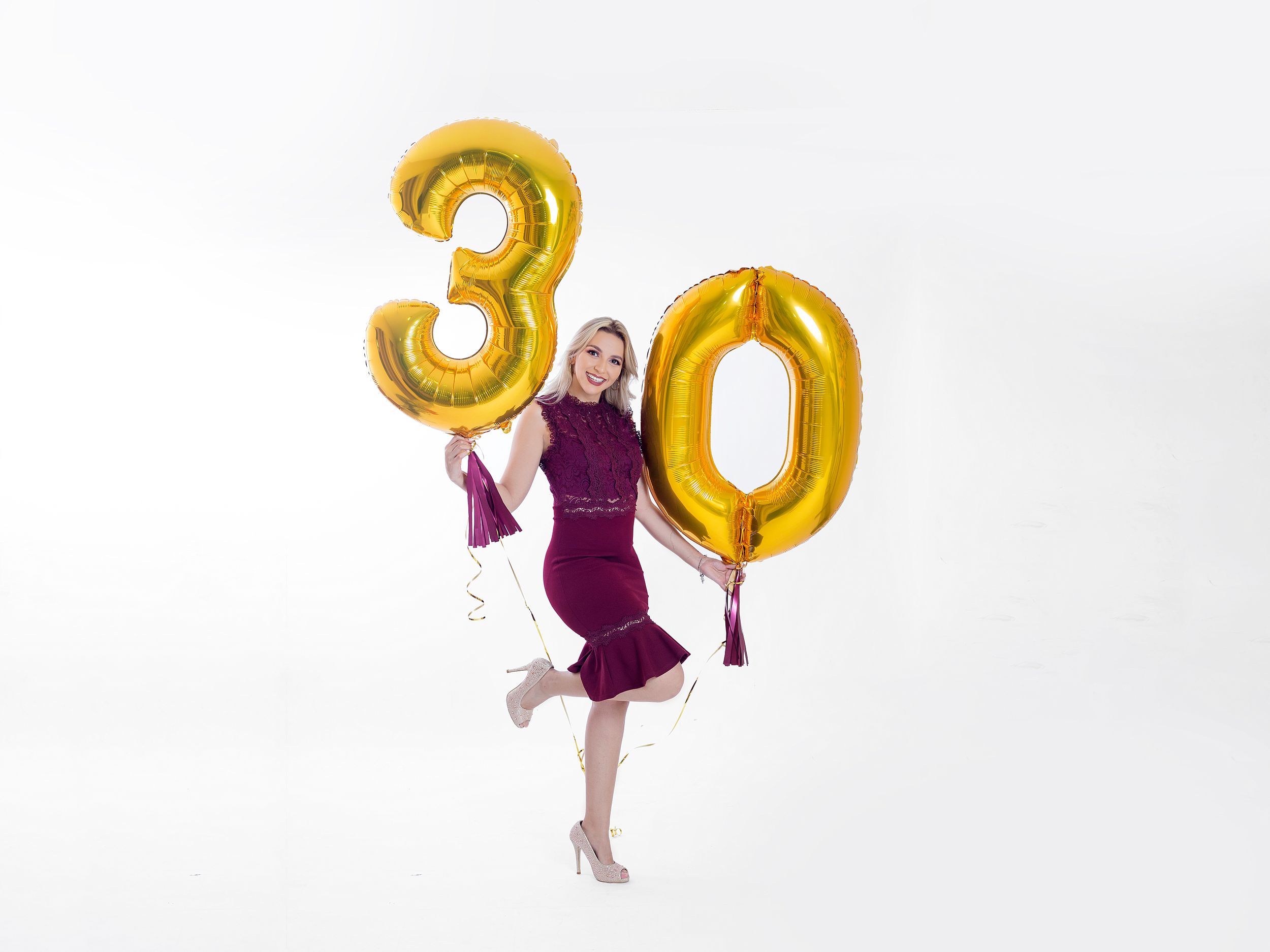 Someone in a purple dress jumps joyfully with gold number 30 balloons against a white background.
