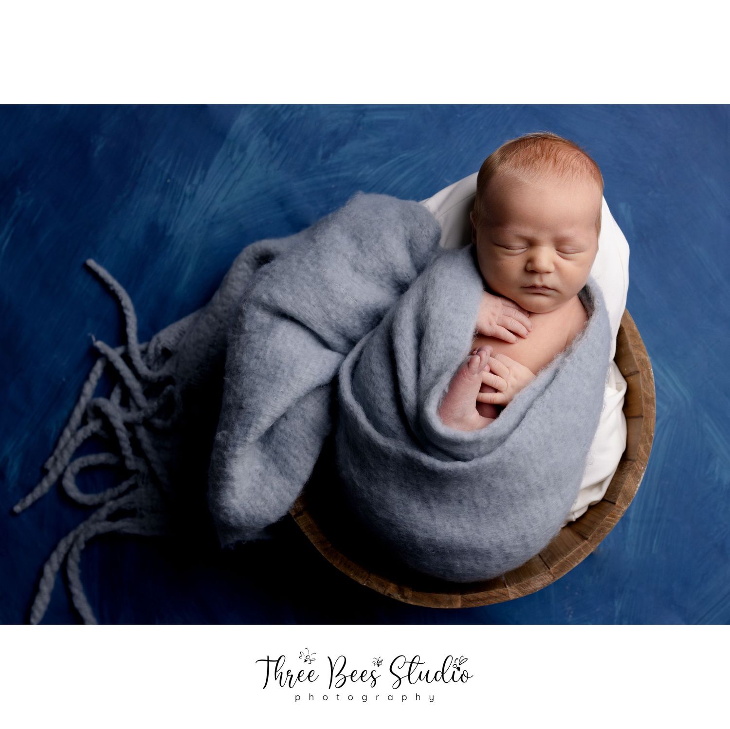 Sleeping newborn wrapped in gray blanket posed in wooden bowl against blue background.