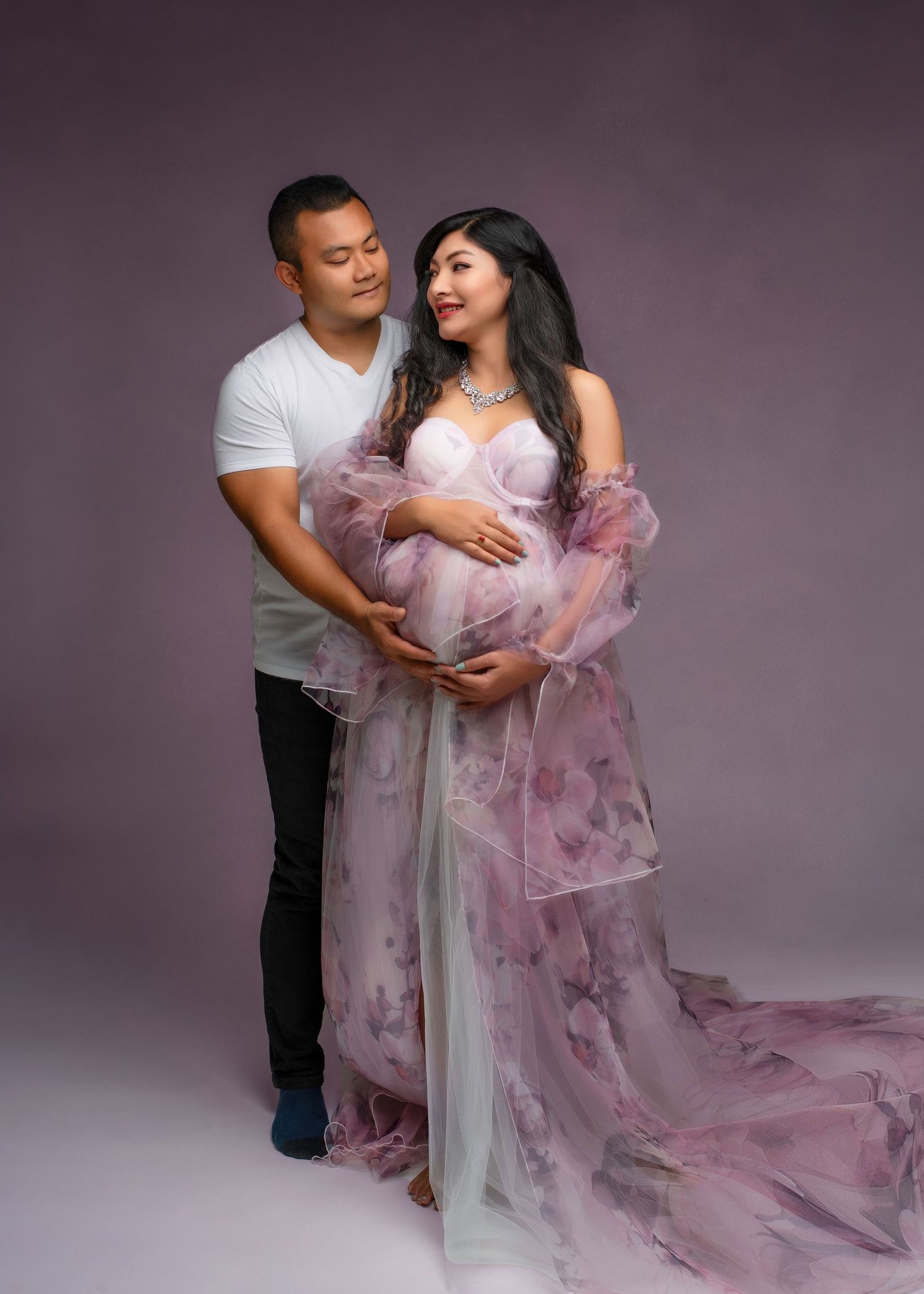 an expecting couple stand together in a photo studio with their hands on her belly