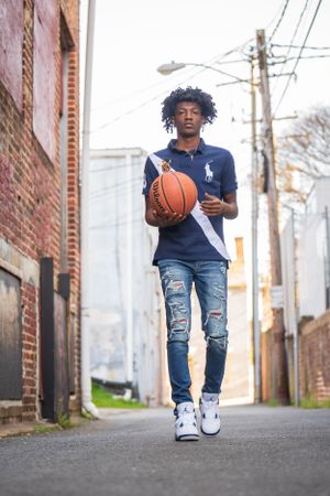 Basketball player walking and dribbling in urban alley with brick buildings and power lines.