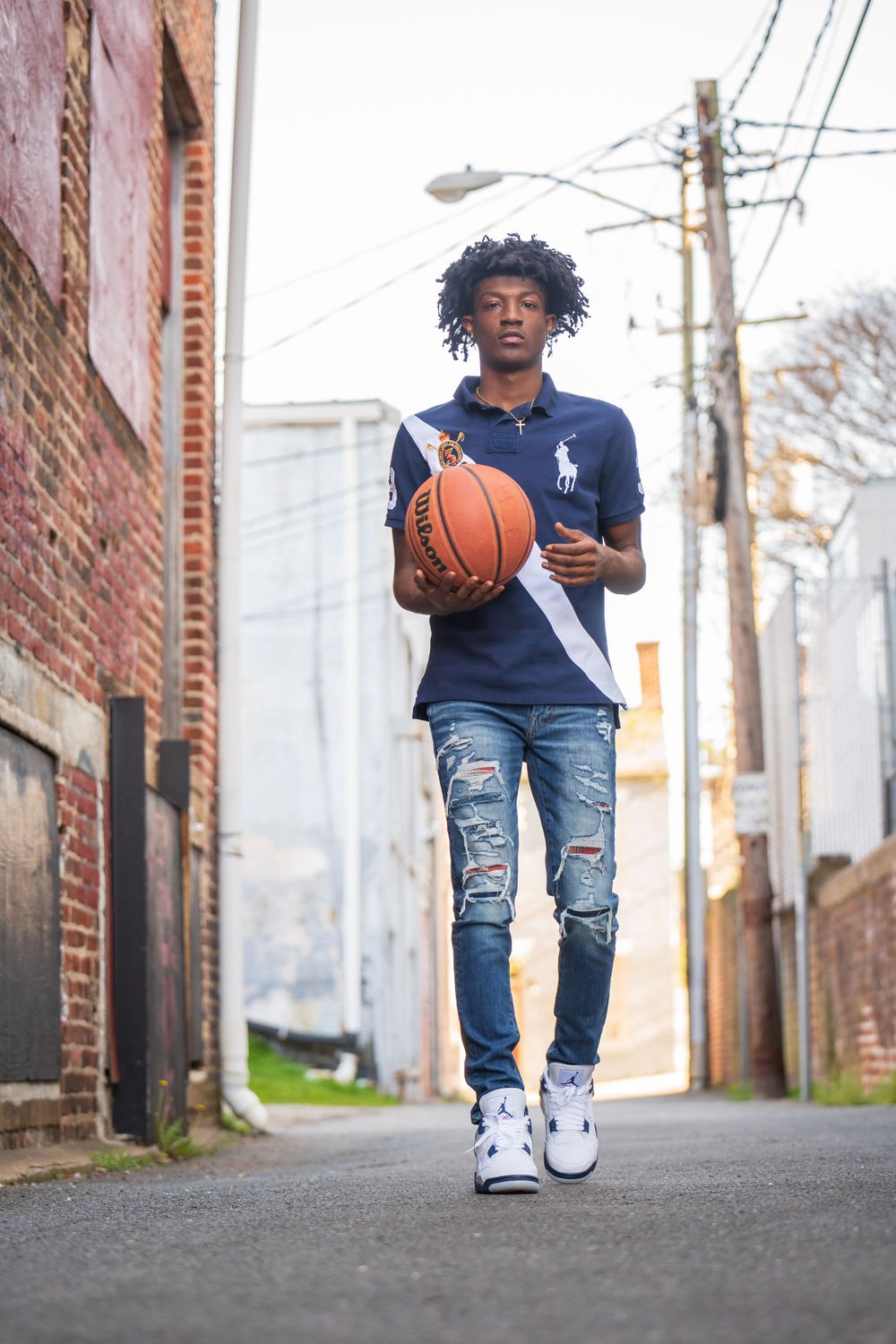 Basketball player walking and dribbling in urban alley with brick buildings and power lines.