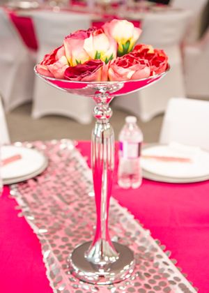 A tall glass centerpiece filled with pink roses sits on a table with silver decor accents.