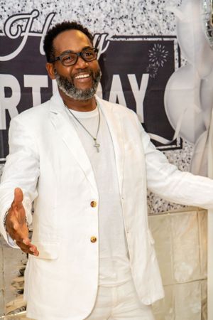 A person in an all-white suit and glasses poses warmly against a birthday celebration backdrop.