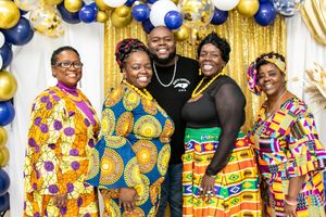 A group poses under blue and gold balloon arch wearing vibrant African print outfits.