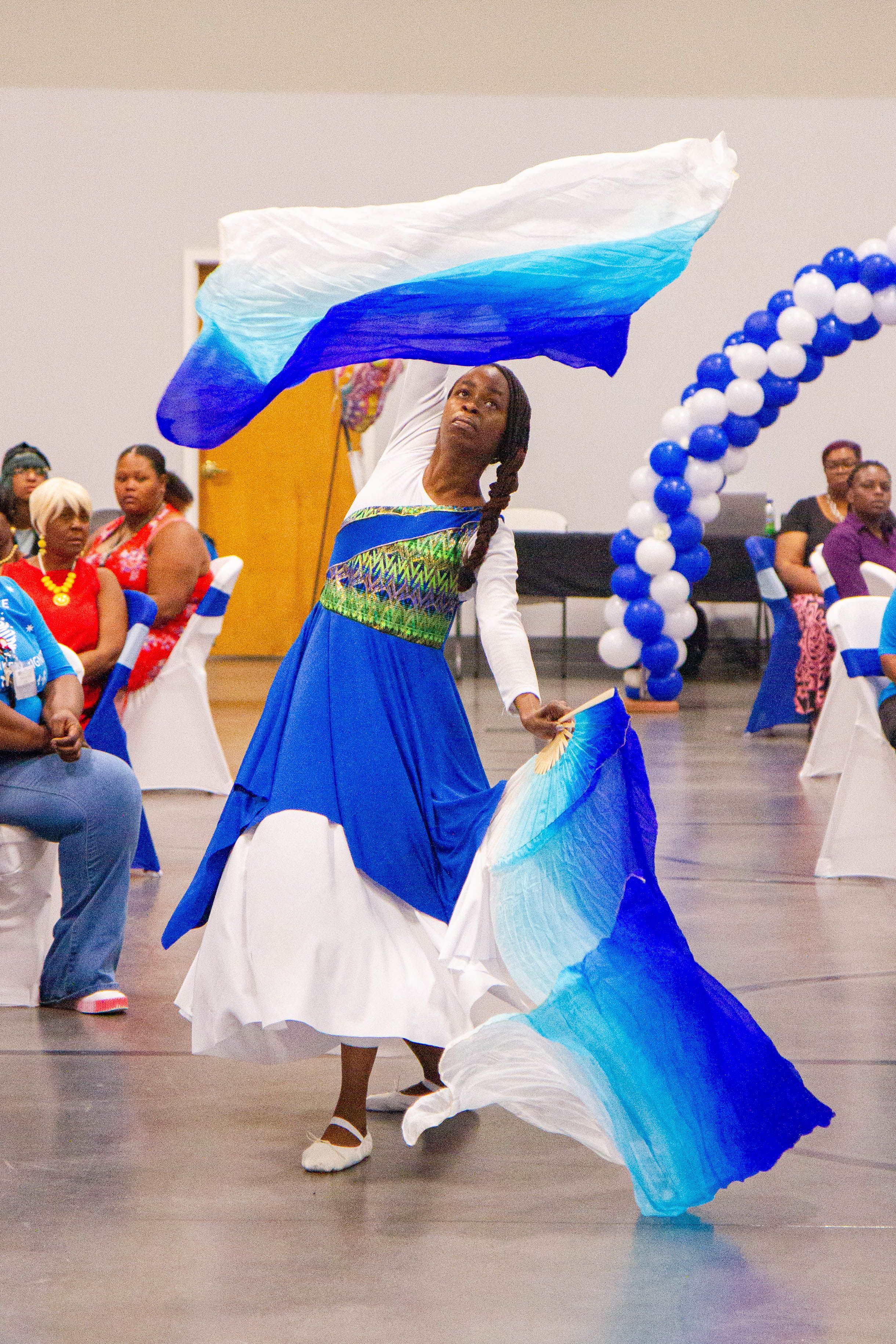 Dancer in flowing blue and white dress performs with ribbons and balloons in the background.