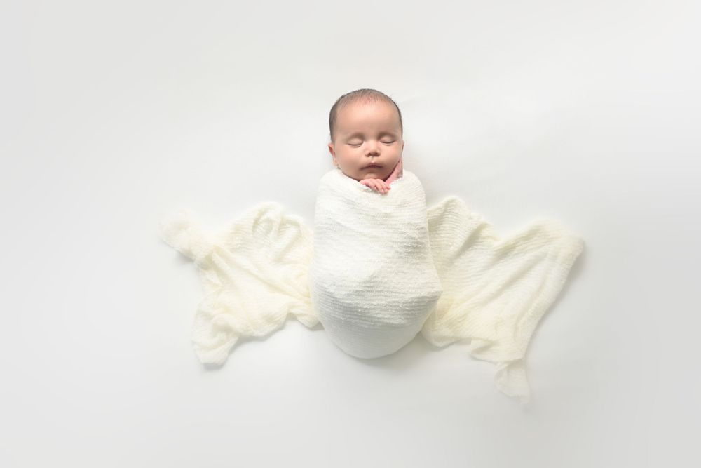 Baby wrapped in white swaddle blanket against plain white background.