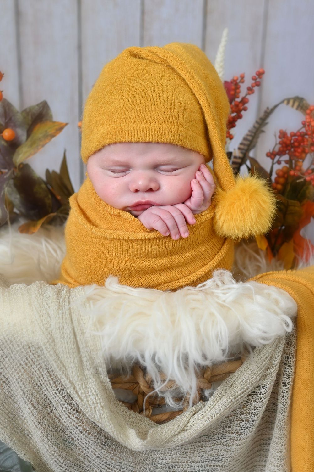 Sleeping newborn in mustard yellow knit wrap with autumn decorations and white fur backdrop.