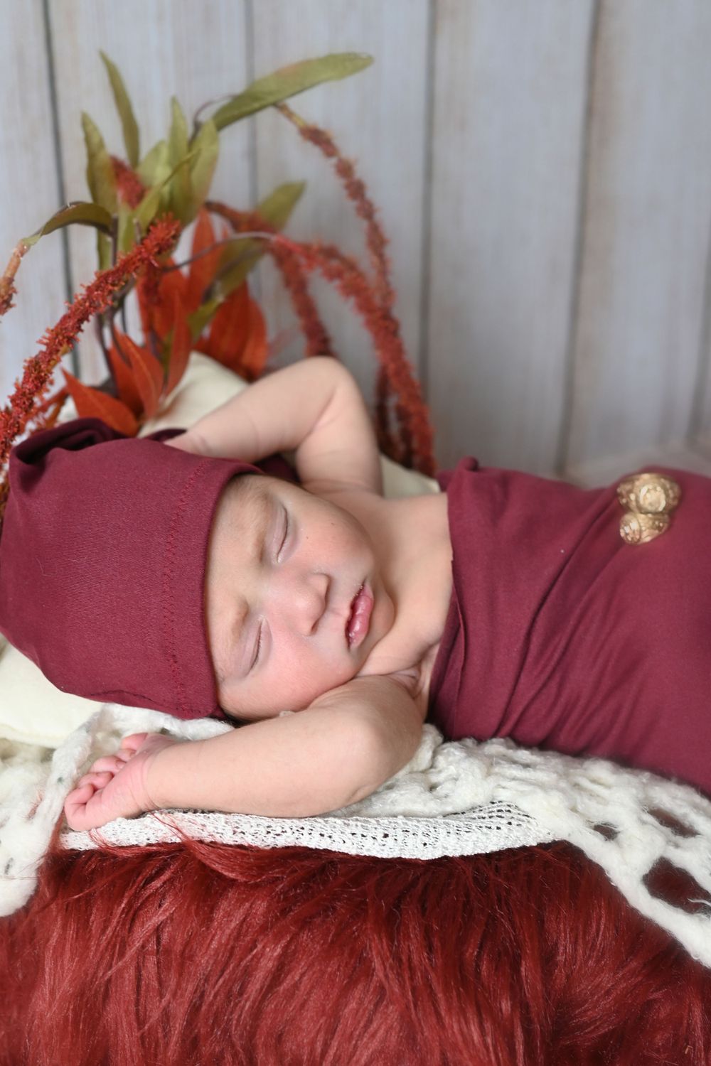 Sleeping newborn in burgundy clothing with orange floral accents and white lace background.
