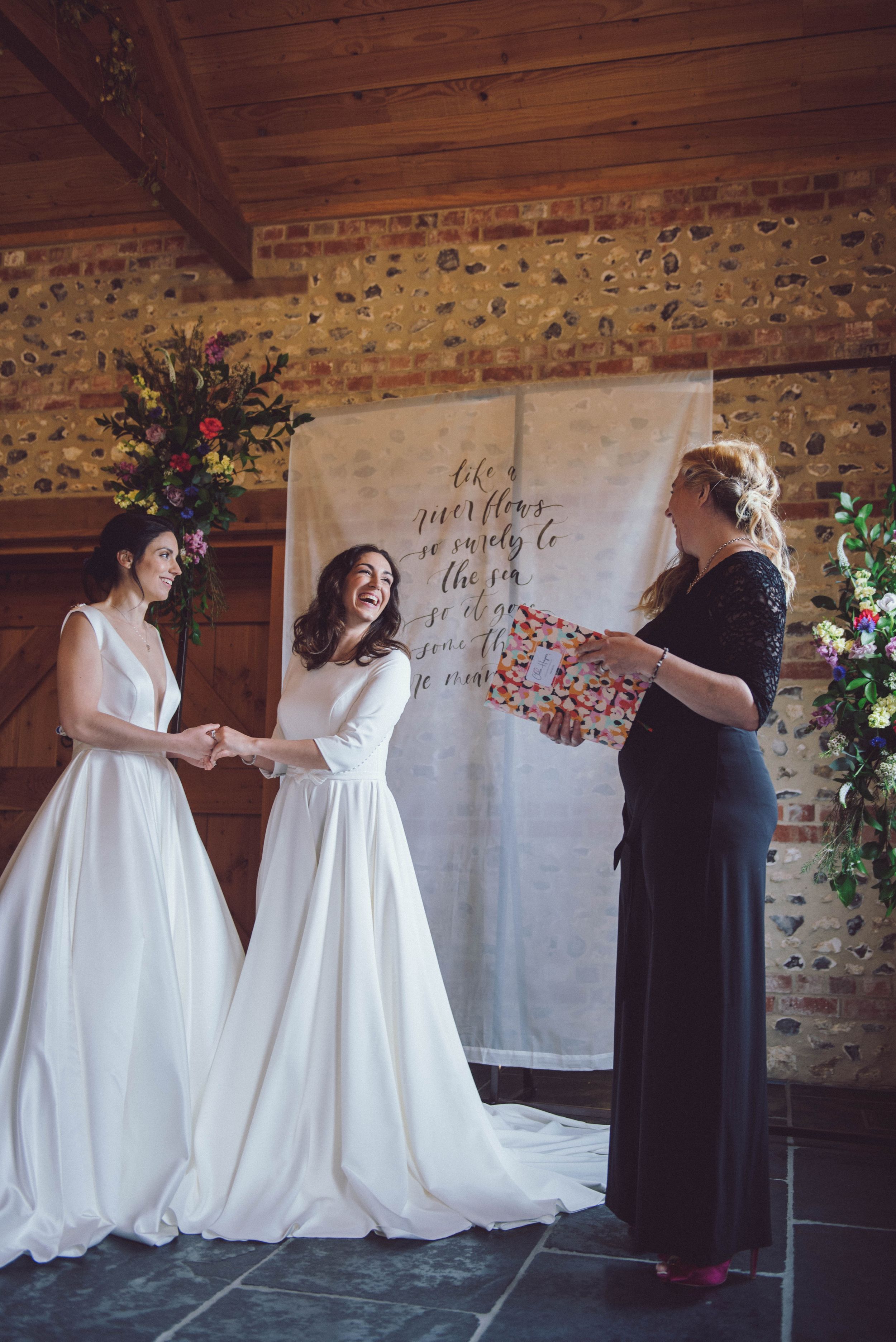 Two people in white dresses stand together against a rustic brick wall with handwritten text.