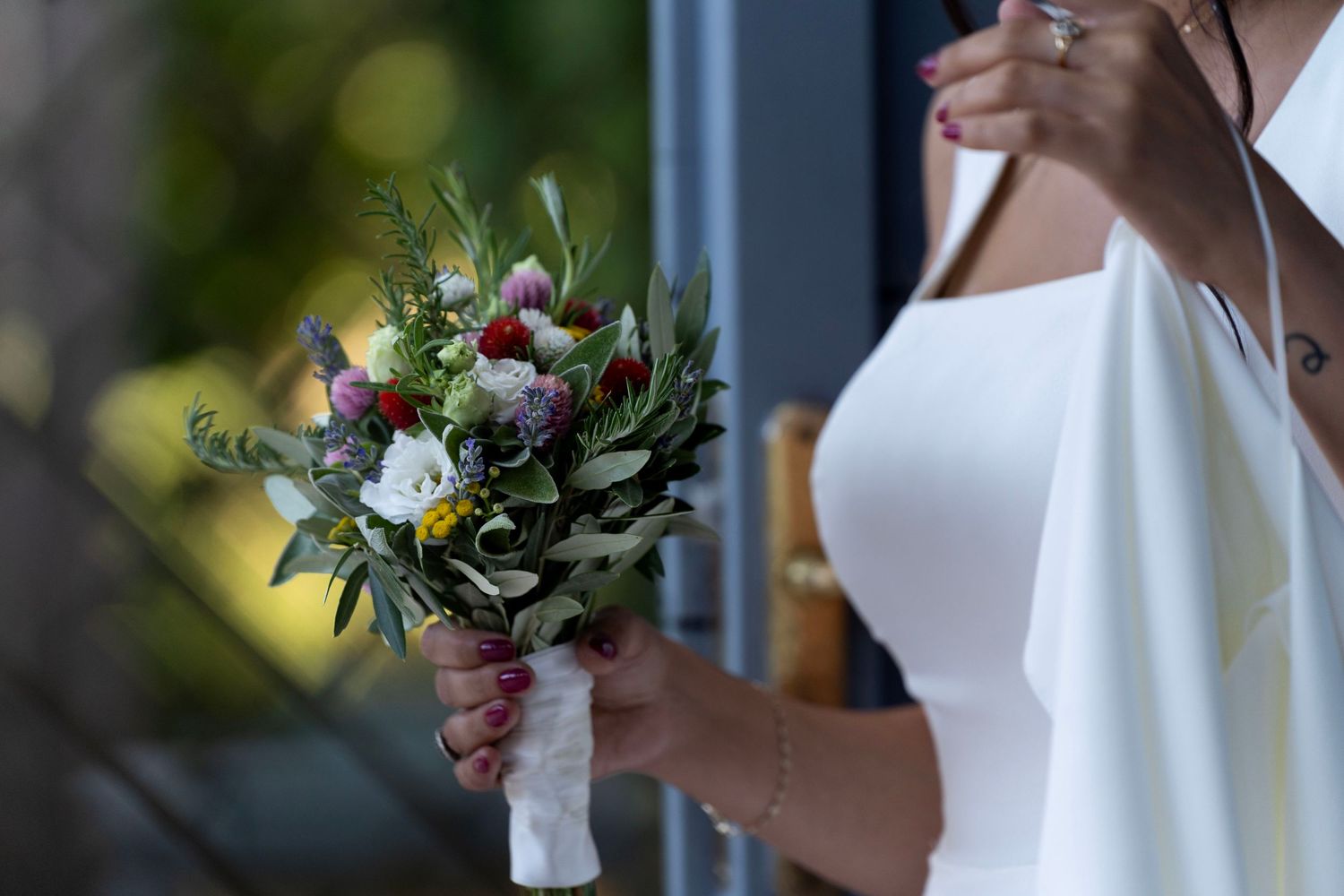 A delicate bouquet of mixed wildflowers and greenery wrapped in white ribbon being held against a light background.