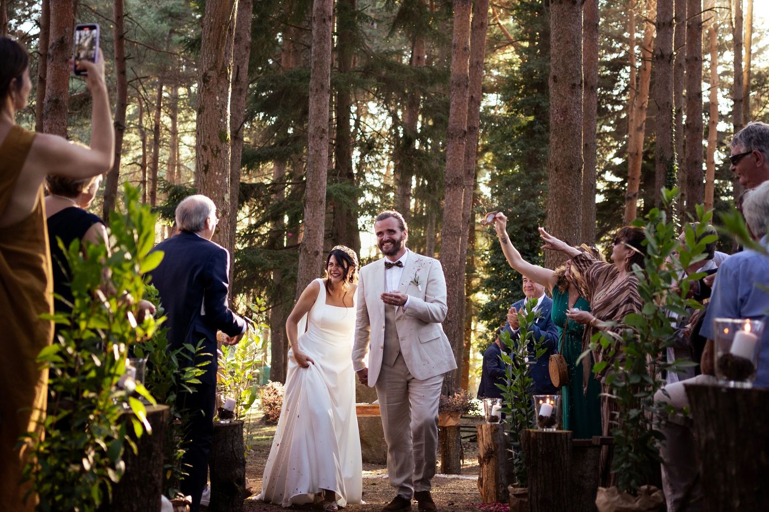 Newlyweds walk down the forest aisle as guests celebrate with raised hands.