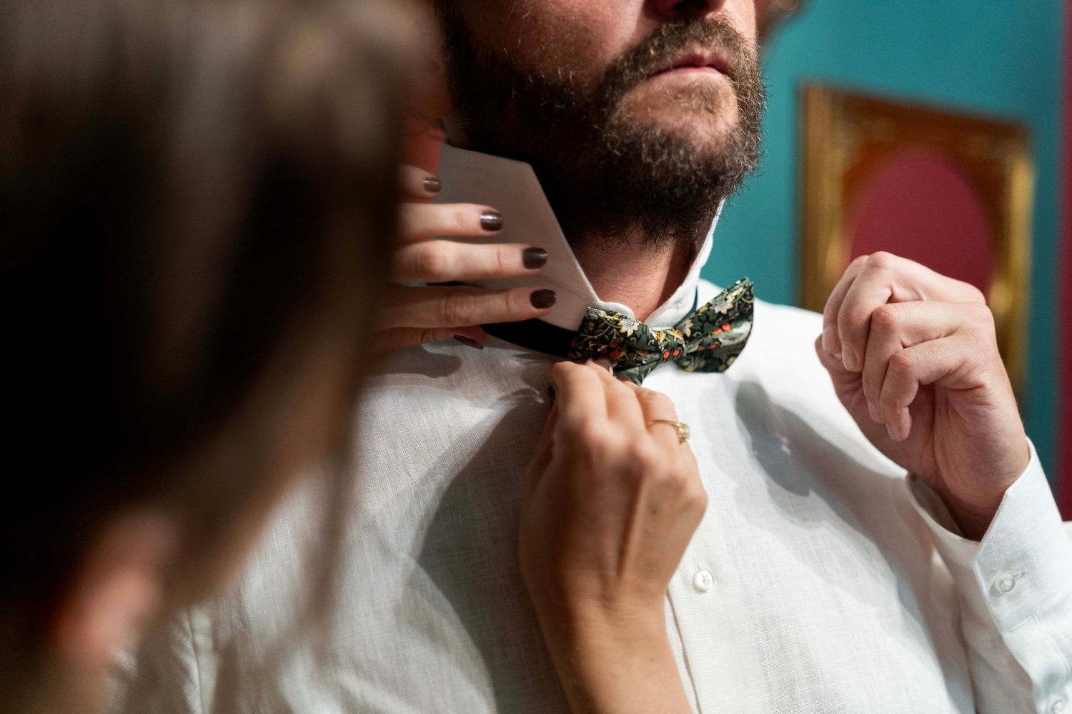 Someone adjusts a floral bowtie on a white dress shirt against a teal background.