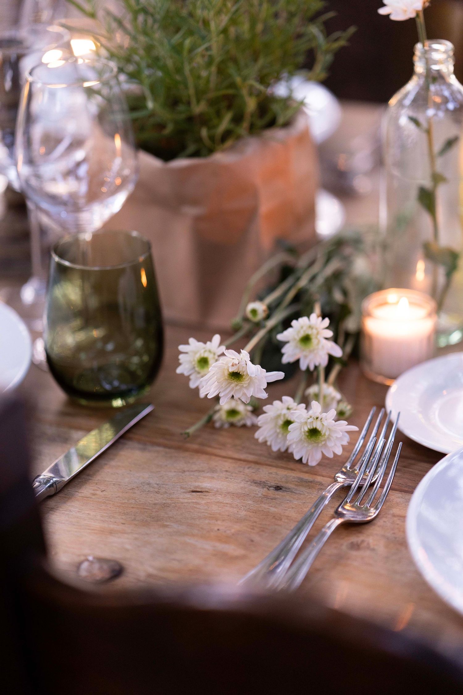 Rustic table setting with white daisies, wine glasses, and candlelight on wooden surface.
