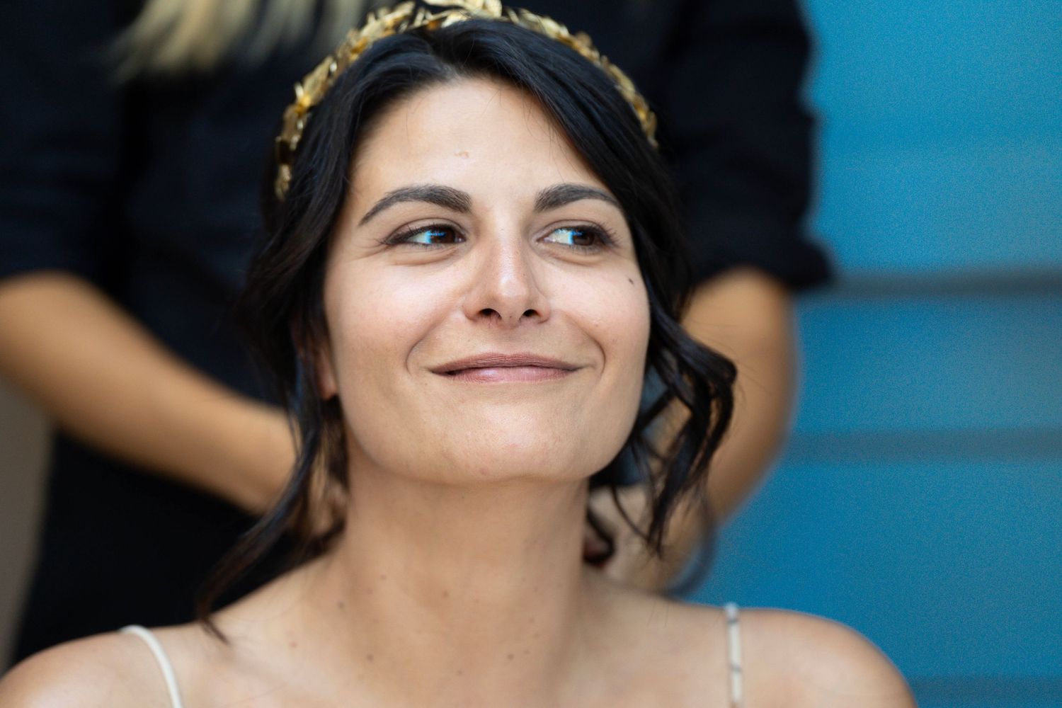 A person wearing a gold headpiece smiles warmly against a blue background.