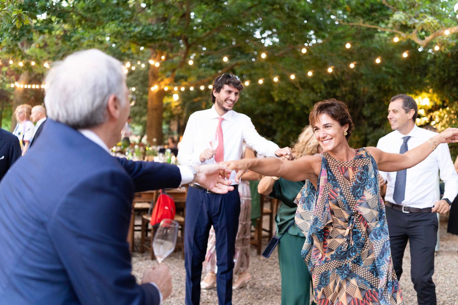 People dancing and celebrating at an outdoor party with string lights in the background.