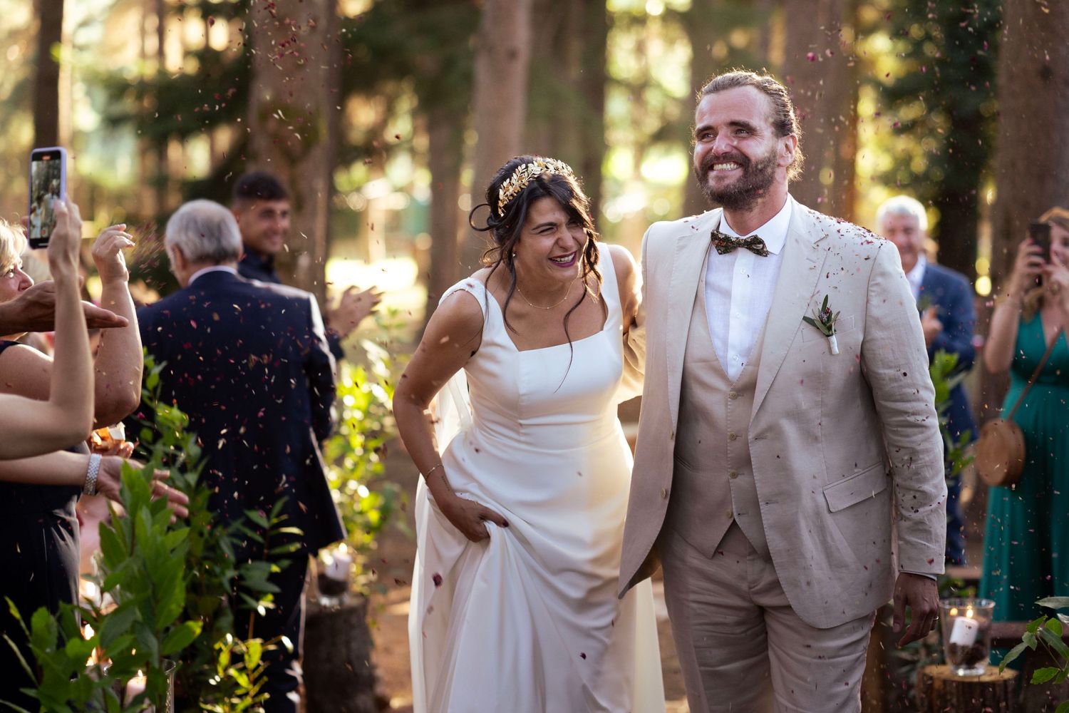 Joyful newlyweds walk through a forest as wedding guests cheer and celebrate.