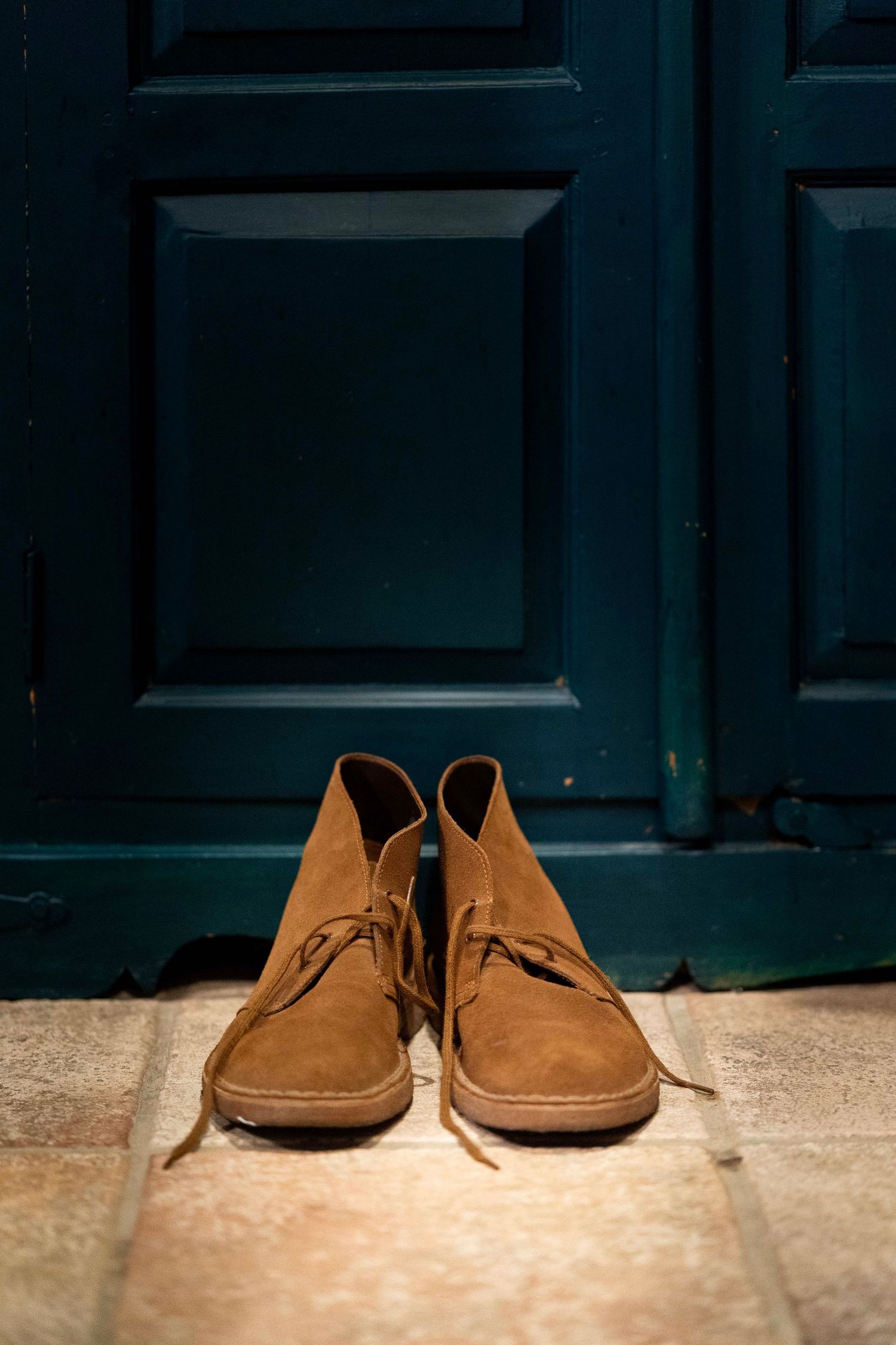 A pair of brown suede desert boots positioned in front of a dark teal door.