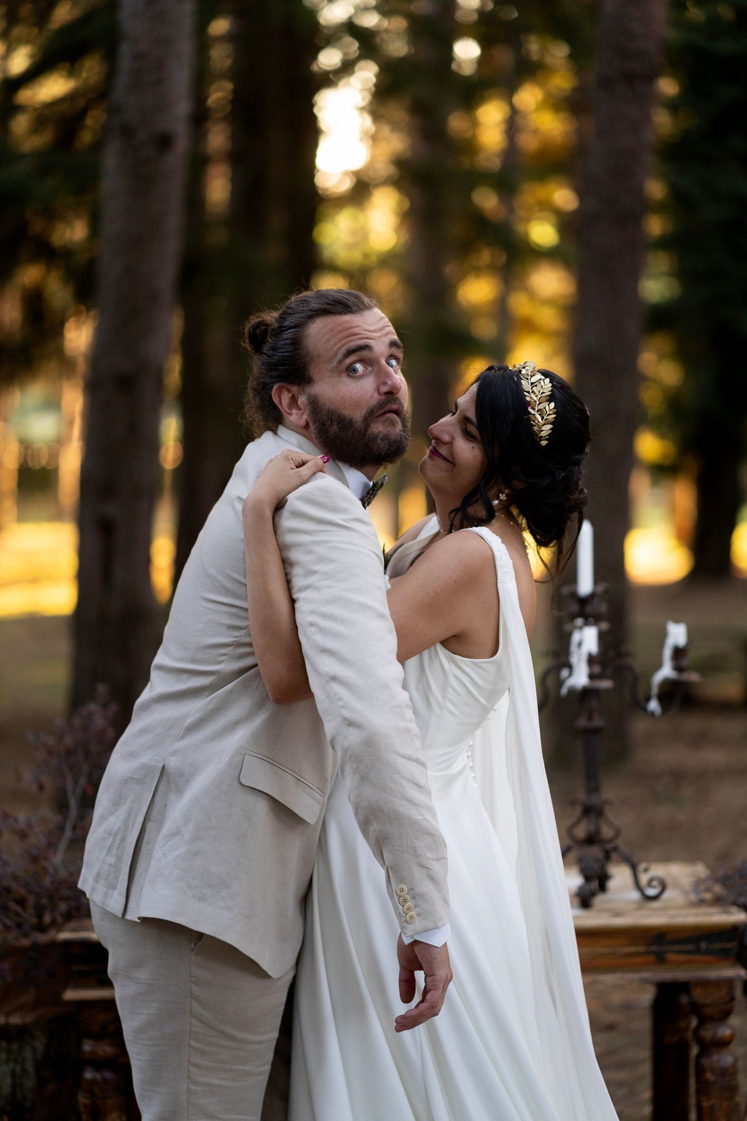 A playful wedding portrait with couple striking a fun pose in front of sunset-lit forest trees.