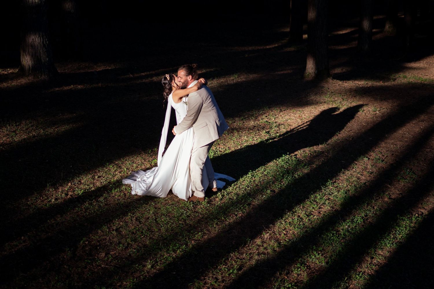 Newlyweds share an intimate moment in a forest with dramatic shadows cast by sunlight through trees.