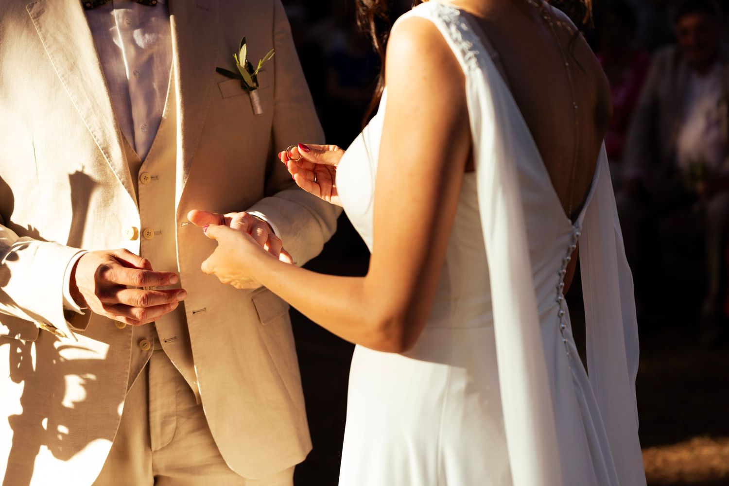 A couple's hands exchanging rings during a sunlit outdoor wedding ceremony.