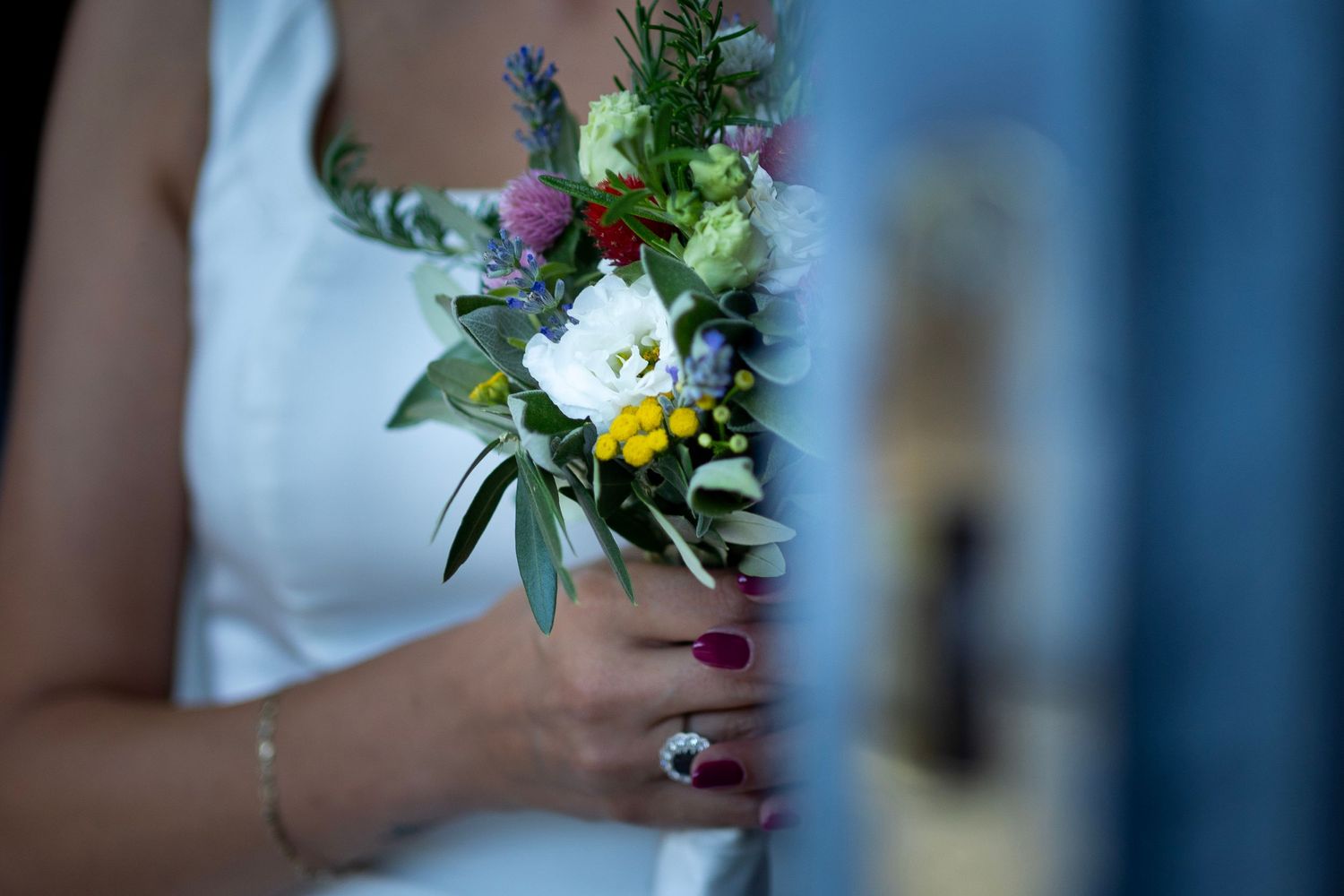 Close-up of a rustic wedding bouquet with yellow and purple wildflowers held by hands in a white dress.
