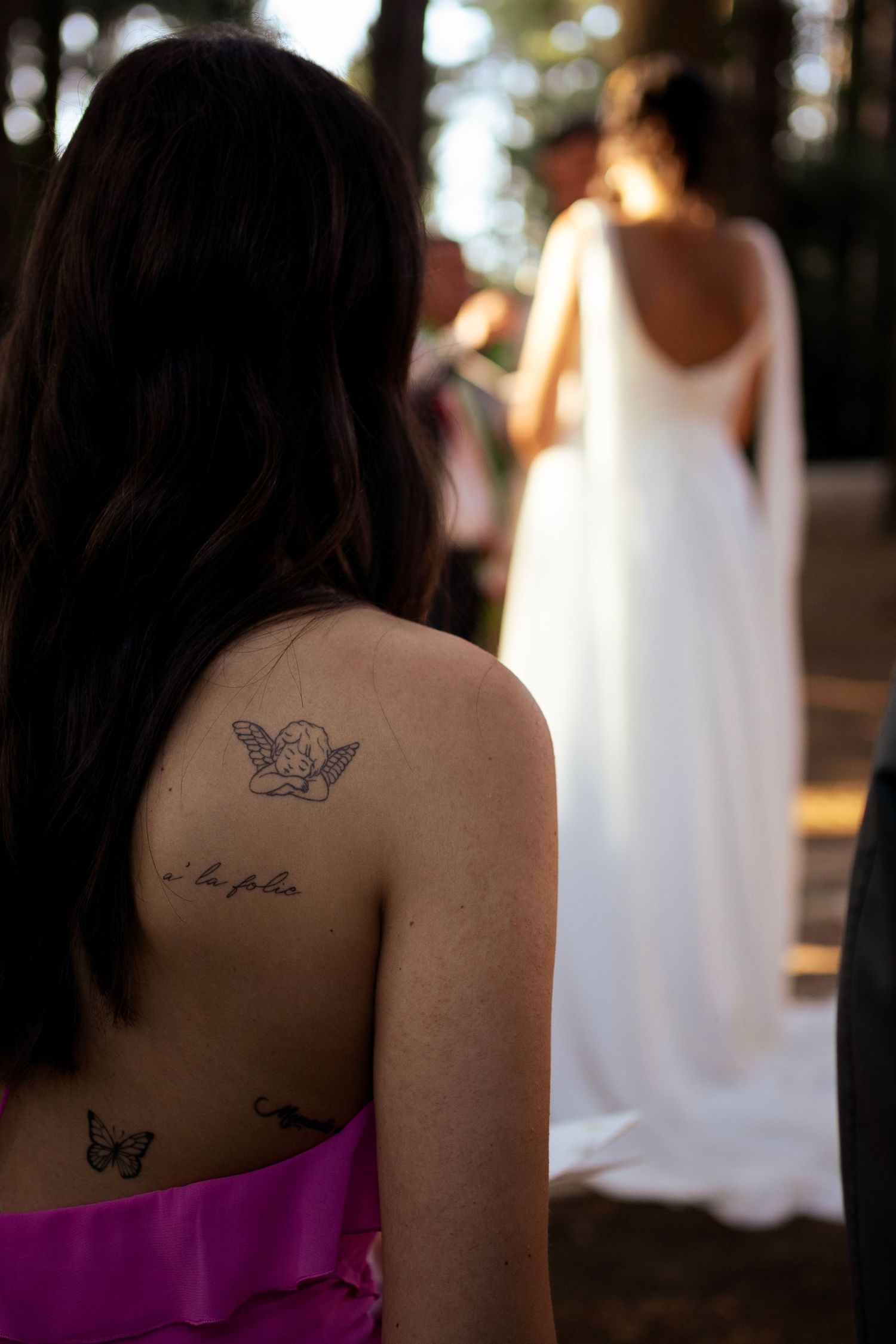 Guest with butterfly tattoos watches wedding ceremony in forest setting.