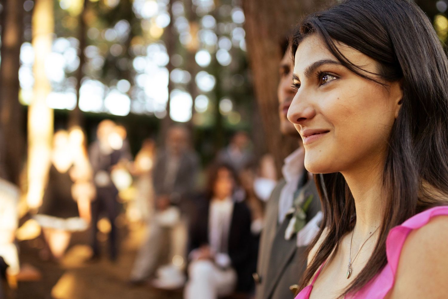 A wedding guest smiles while attending an outdoor forest celebration.