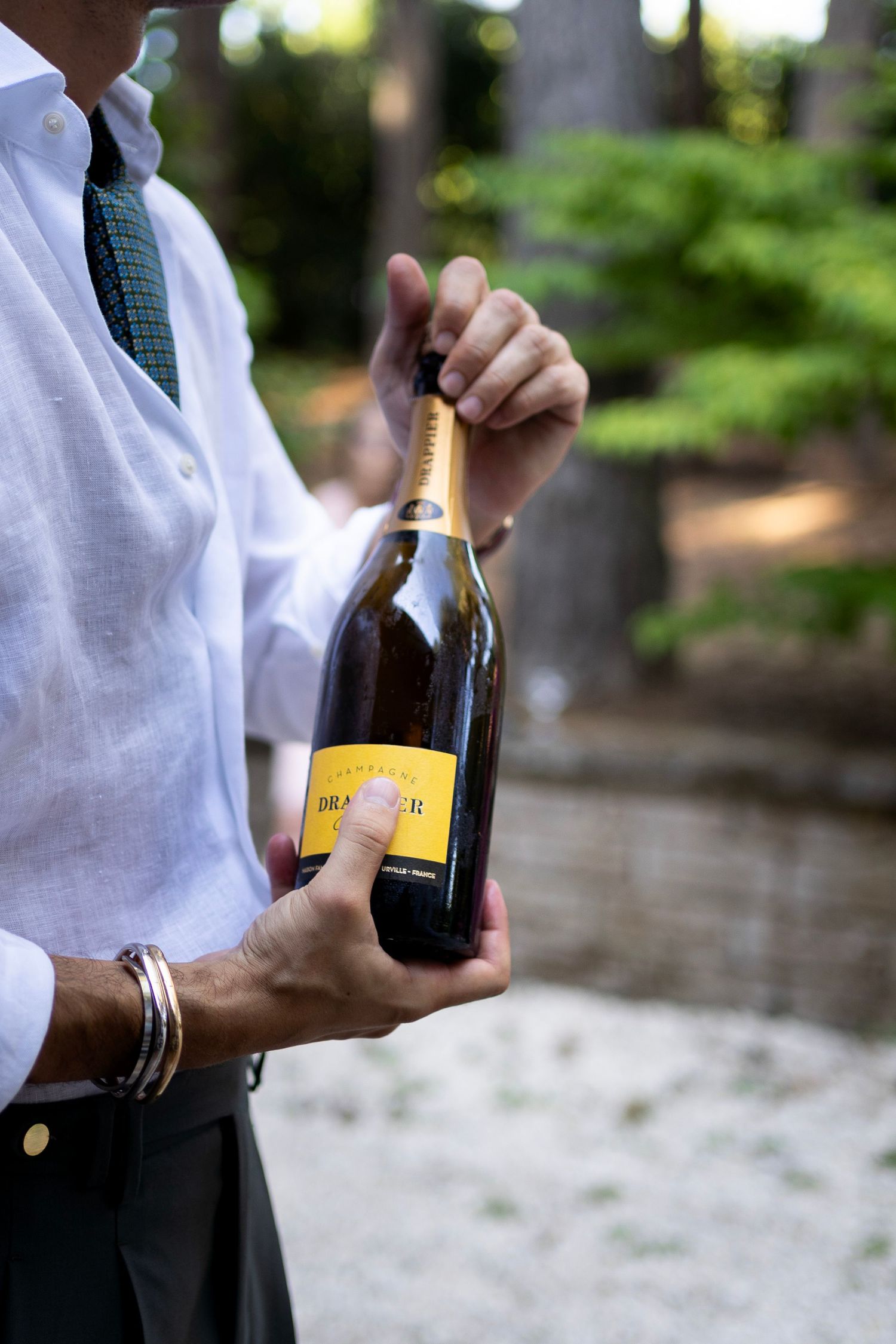 Hands hold a bottle of champagne while wearing a white shirt and green tie outdoors.