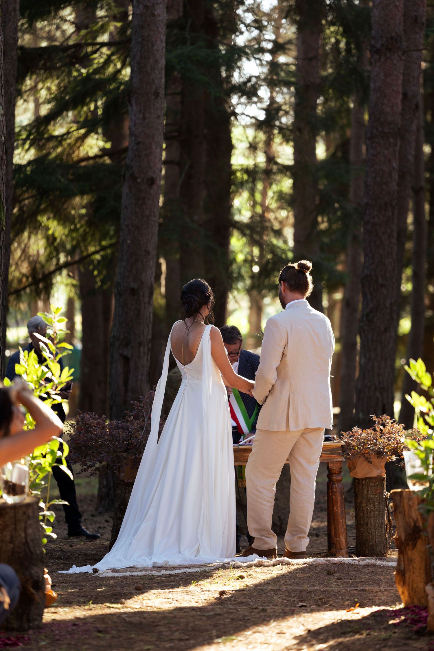 Couple faces an officiant during their forest wedding ceremony with sunlight filtering through trees.